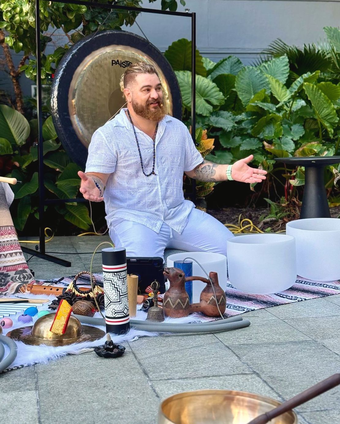 Juan Saer in white, kneeling in front of a gong and speaking with open hands, his instruments laid out before him